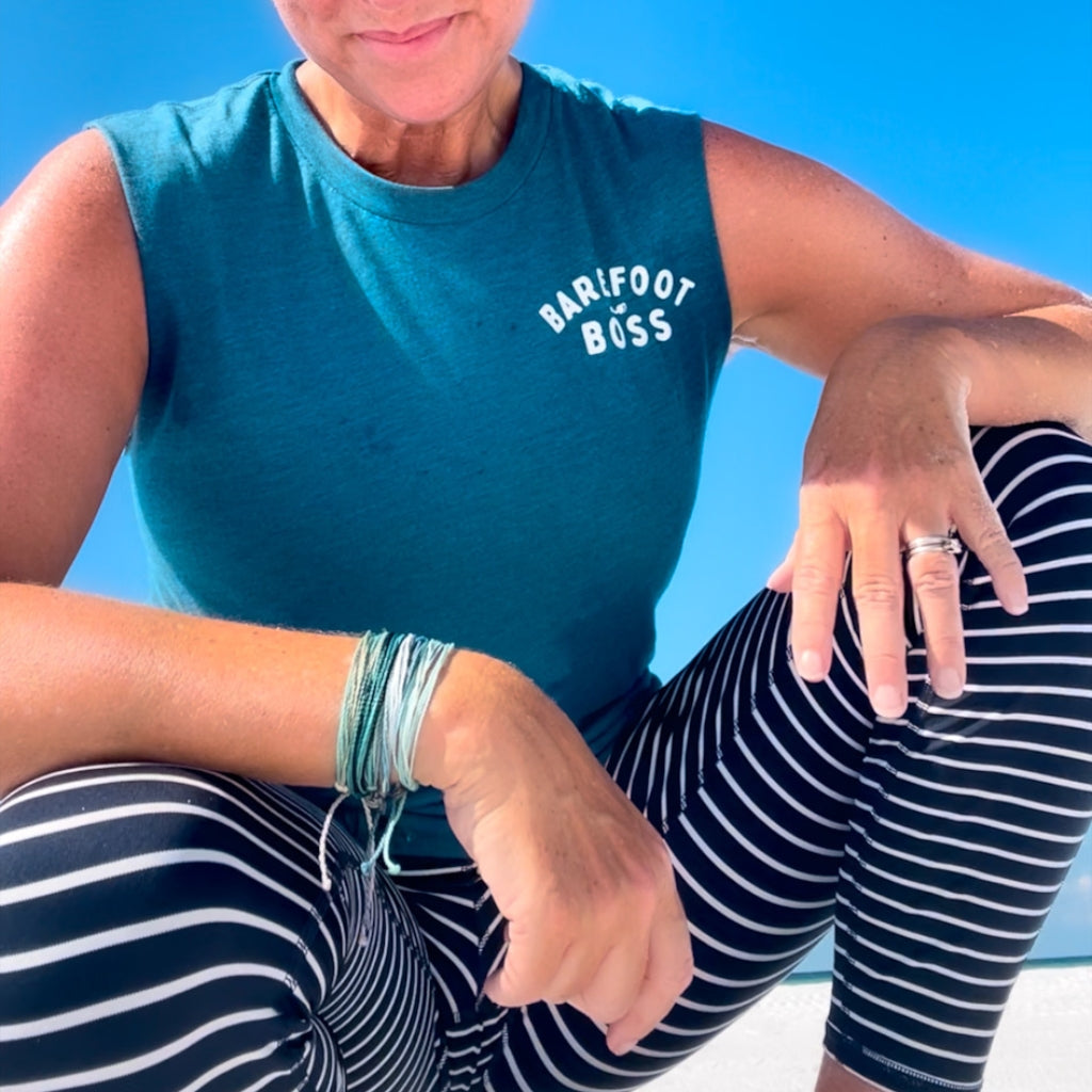 Person wearing a teal sleeveless shirt with 'Barfoot Boss' text and striped pants on the sandy beach.