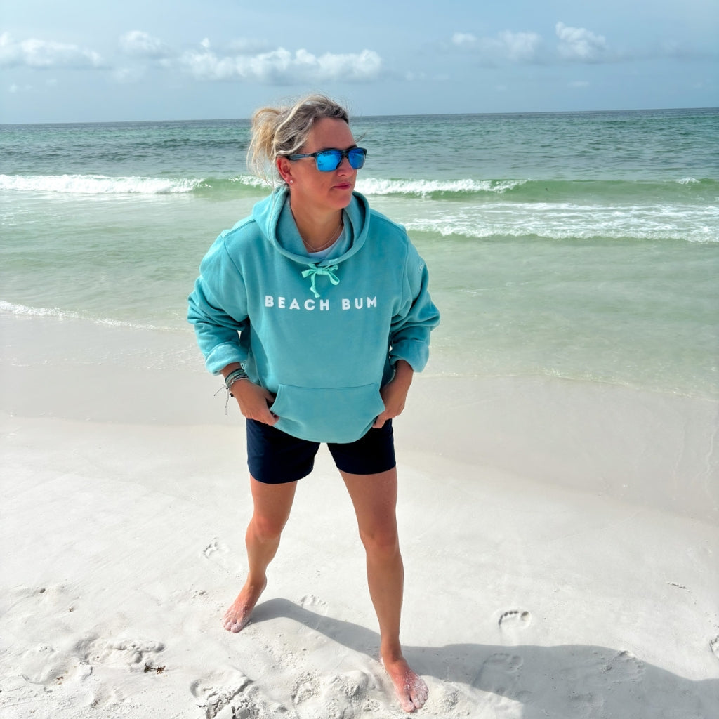 Person wearing a teal 'Beach Bum' hoodie on a sandy beach with ocean in the background.  Beach apparel.  Beachwear.  Beach Bum hoodie.