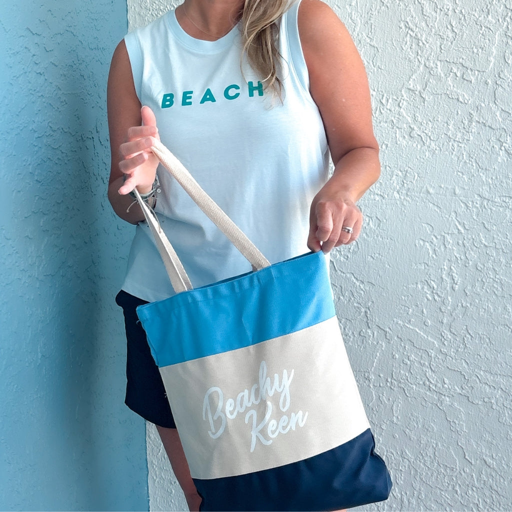 Person holding a blue and white tote bag with 'Beach' text on a light blue wall.  Beachy Keen tote.  Beach tote.