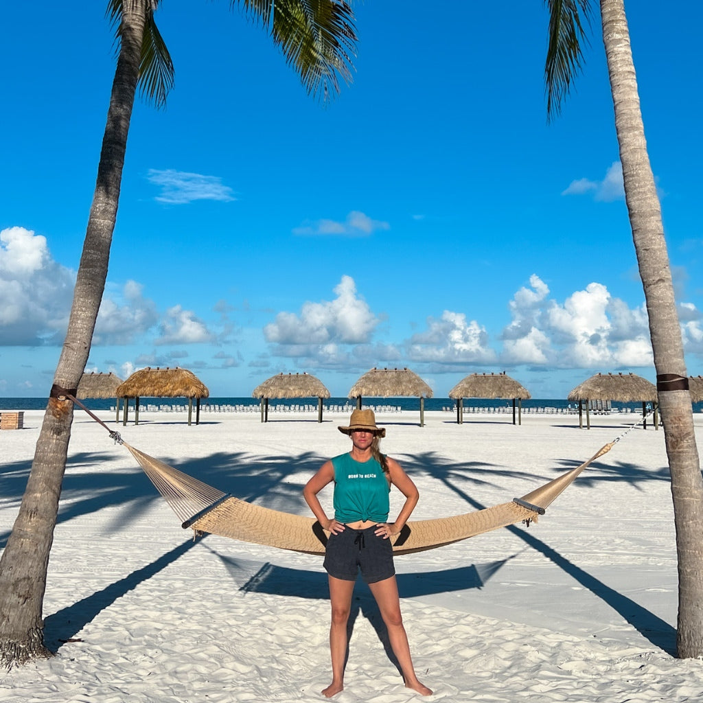 born to beach white text on raw cut unisex muscle tank.  turquoise green colored super soft beach tank.  beach apparel.  woman in the sun under palm trees near the ocean.