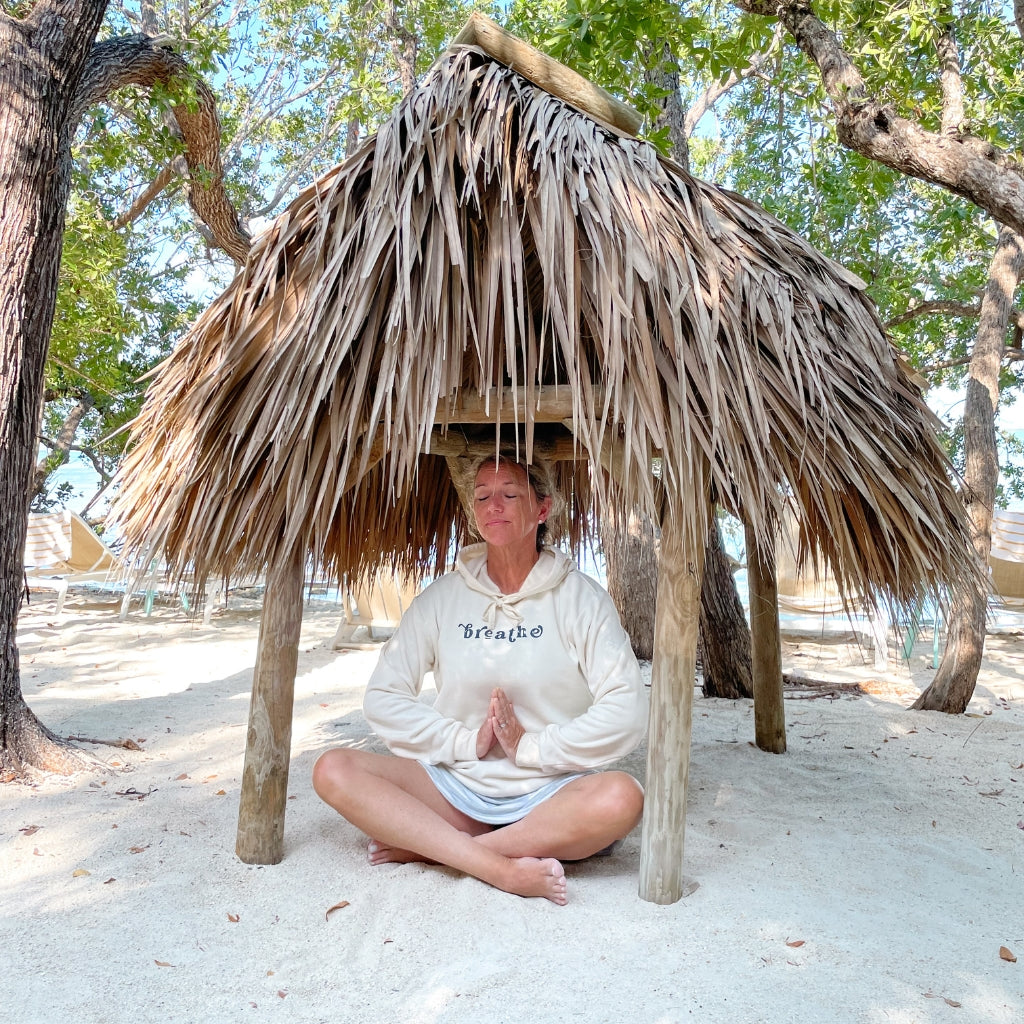 breathe text saying in gray print on an off white unisex hoodie.  breathe.  wellness apparel.  woman wearing a breathe hooded sweatshirt sitting under a palm hut near the ocean.  meditating with a breathe hoodie on.