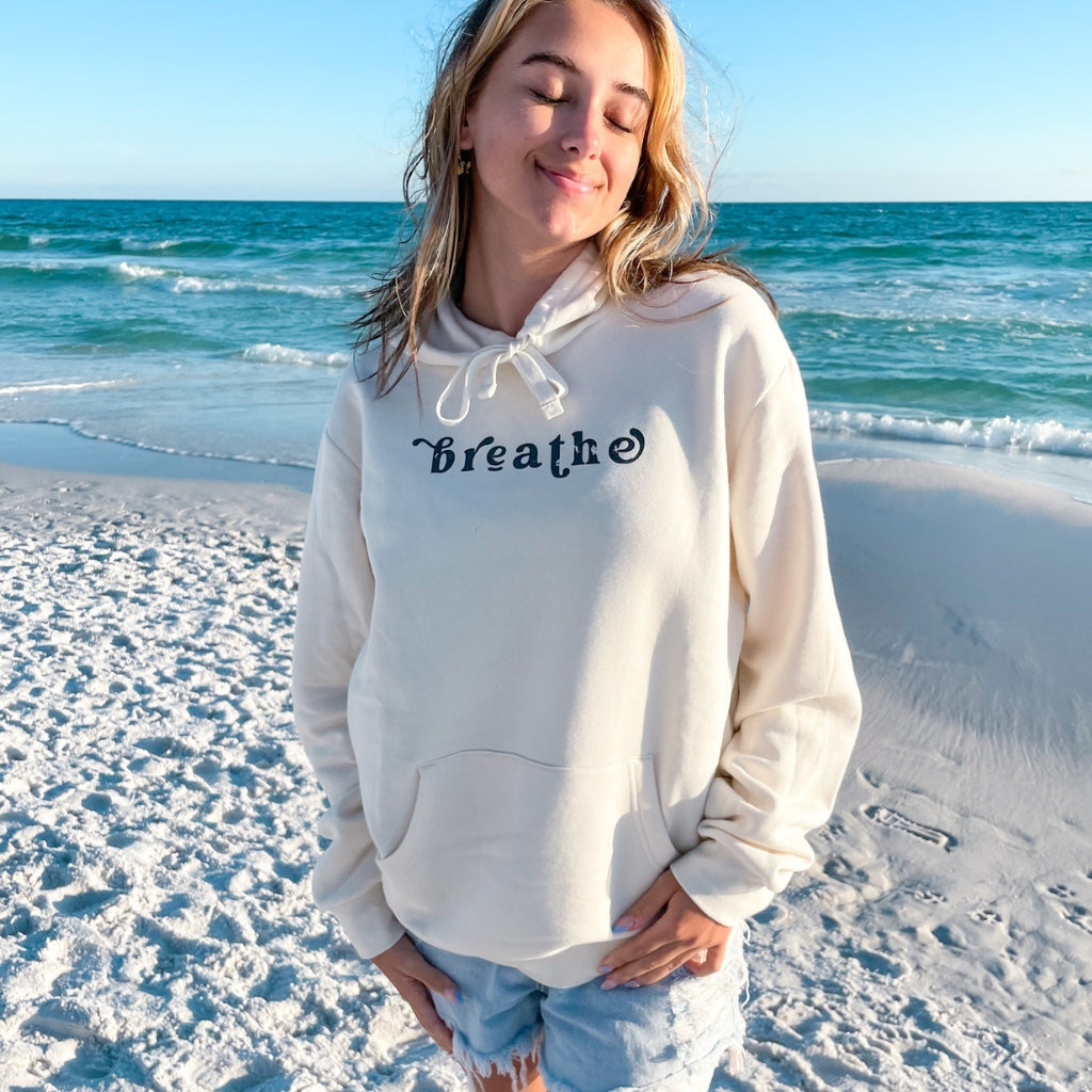 breathe text saying in gray print on an off white unisex hoodie.  breathe.  wellness apparel.  woman wearing a breathe hooded sweatshirt near the ocean.