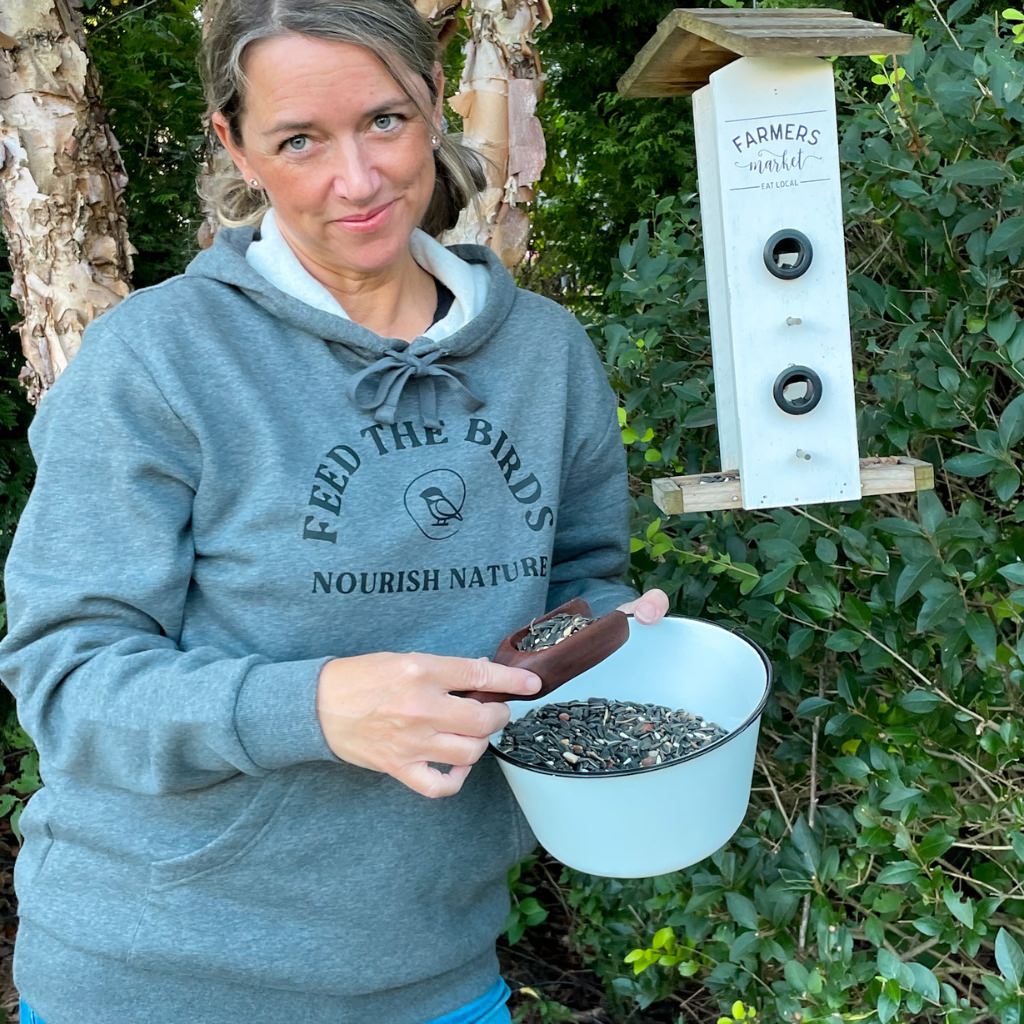 Woman feeding the birds wearing her relaxed super soft gray hoodie with text saying Feed the Birds, Nourish Nature.  Unisex sized hoodie.  chickadee co. bird logo is central on the front of the print design.