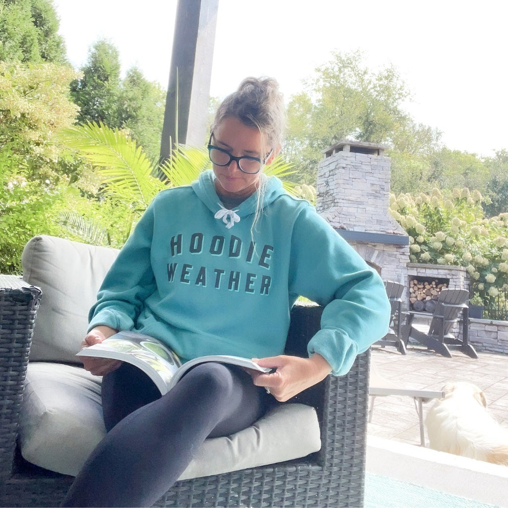 hoodie weather bold printed text on super soft blue hoodie.  text color is two tone black & light blue.  unisex hoodie with saying hoodie weather.  woman wearing soft blue hoodie outdoors on the patio.