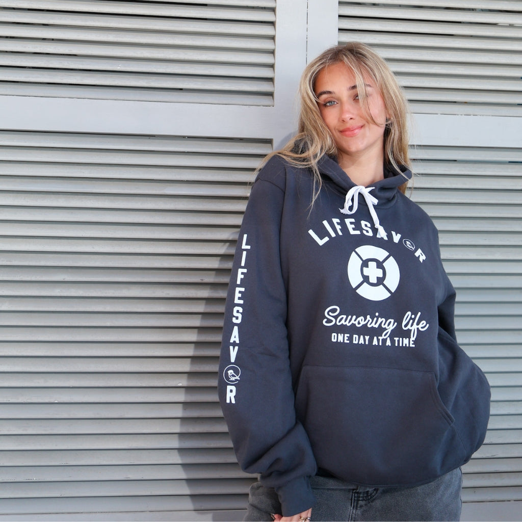 Person wearing a charcoal gray hoodie with 'Lifesavor' on the front and 'Savoring Life' text down the right arm sleeve.
