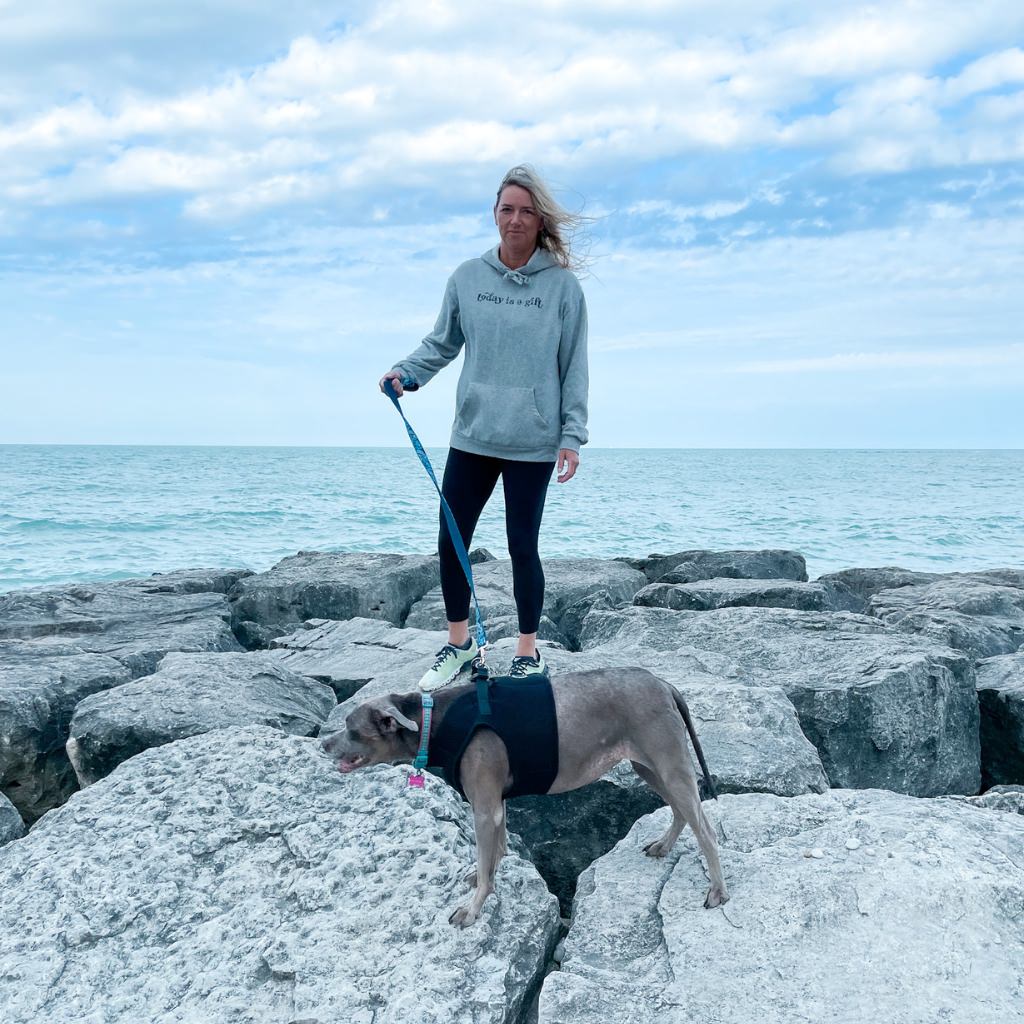 today is a gift, printed in charcoal gray ink on a heathered light gray hoodie.  unisex hoodie with screen printed image text saying today is a gift.  woman standing near a lake with her dog wearing uplifting hoodie.