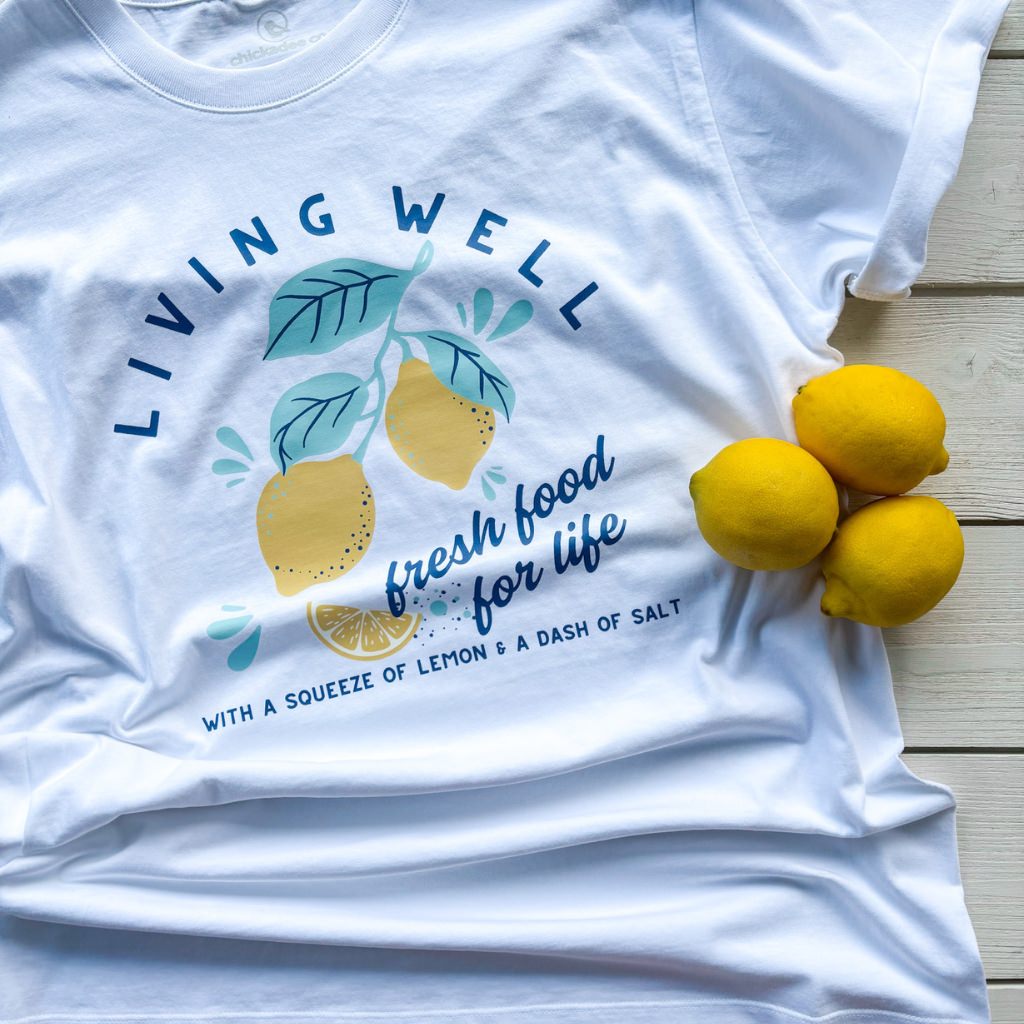 White t-shirt with lemon design and text on a wooden surface.  Living intentionally.  Living well.  Lemon Tshirt.  Organic cotton apparel.  Organic cotton tshirt.