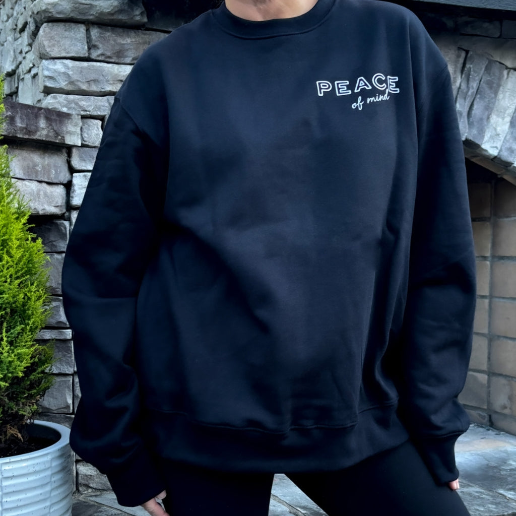 person wearing solid black crew heavyweight sweatshirt with white text 'peace of mind' on front left chest. 'peace of mind' bird graphic & text on full back print.