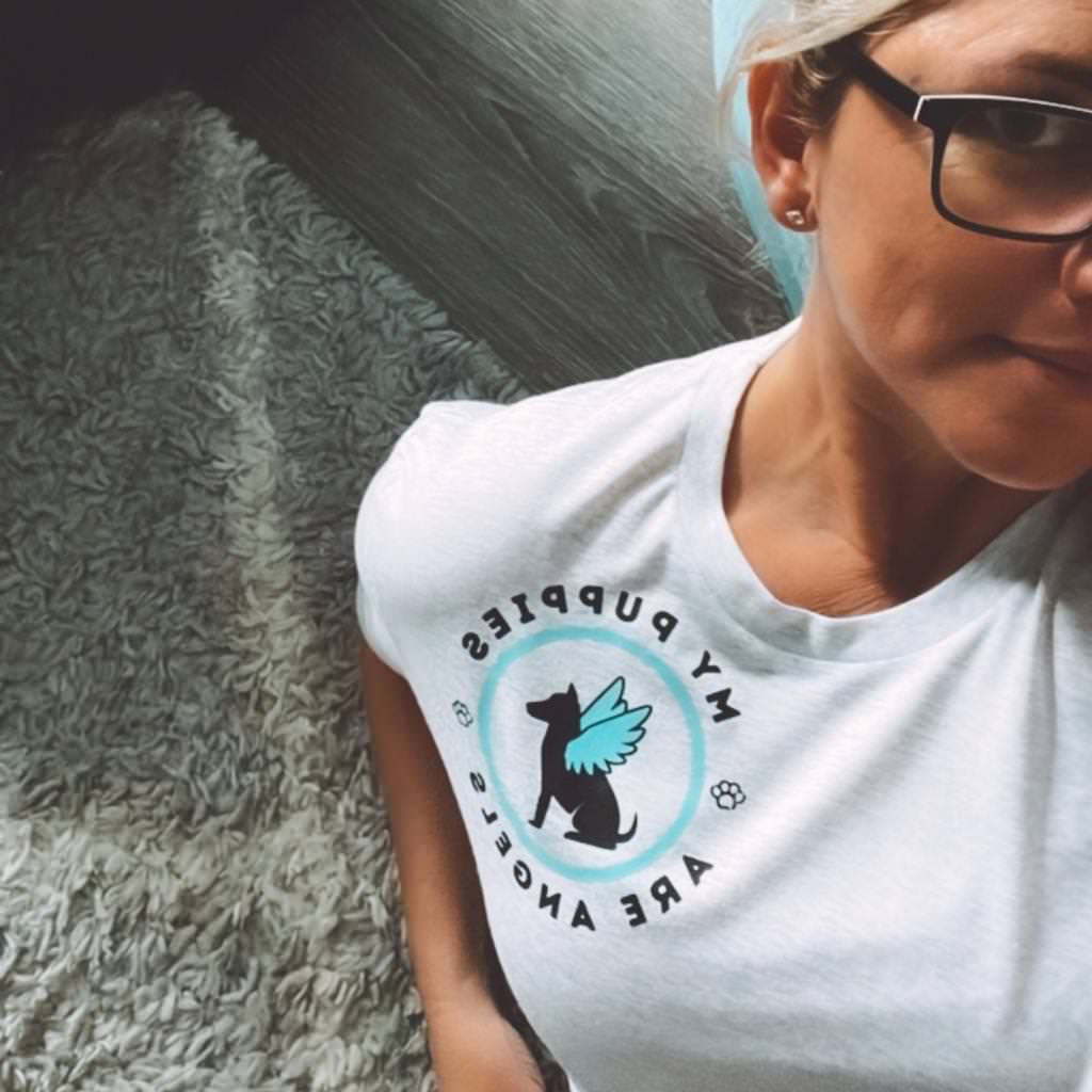 woman wearing a light salty white colored tshirt with upper left print saying 'my puppies are angels'.  circular design with dog and angel wing graphic in blue & black ink.  unisex sized vintage soft dog tshirt.