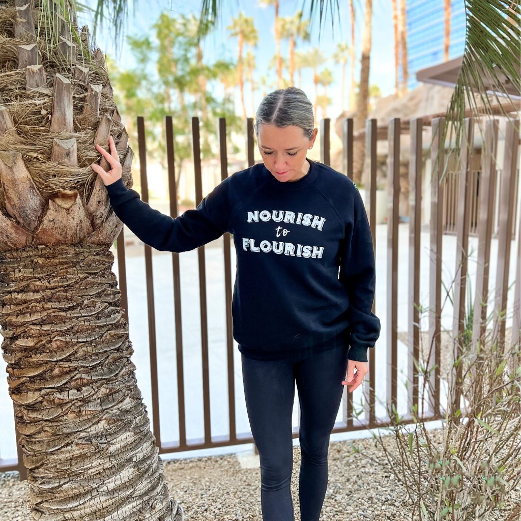 nourish to flourish black unisex crewneck sweatshirt.  super soft wellness apparel.  woman wearing black sweatshirt with white ink saying ' nourish to flourish'.