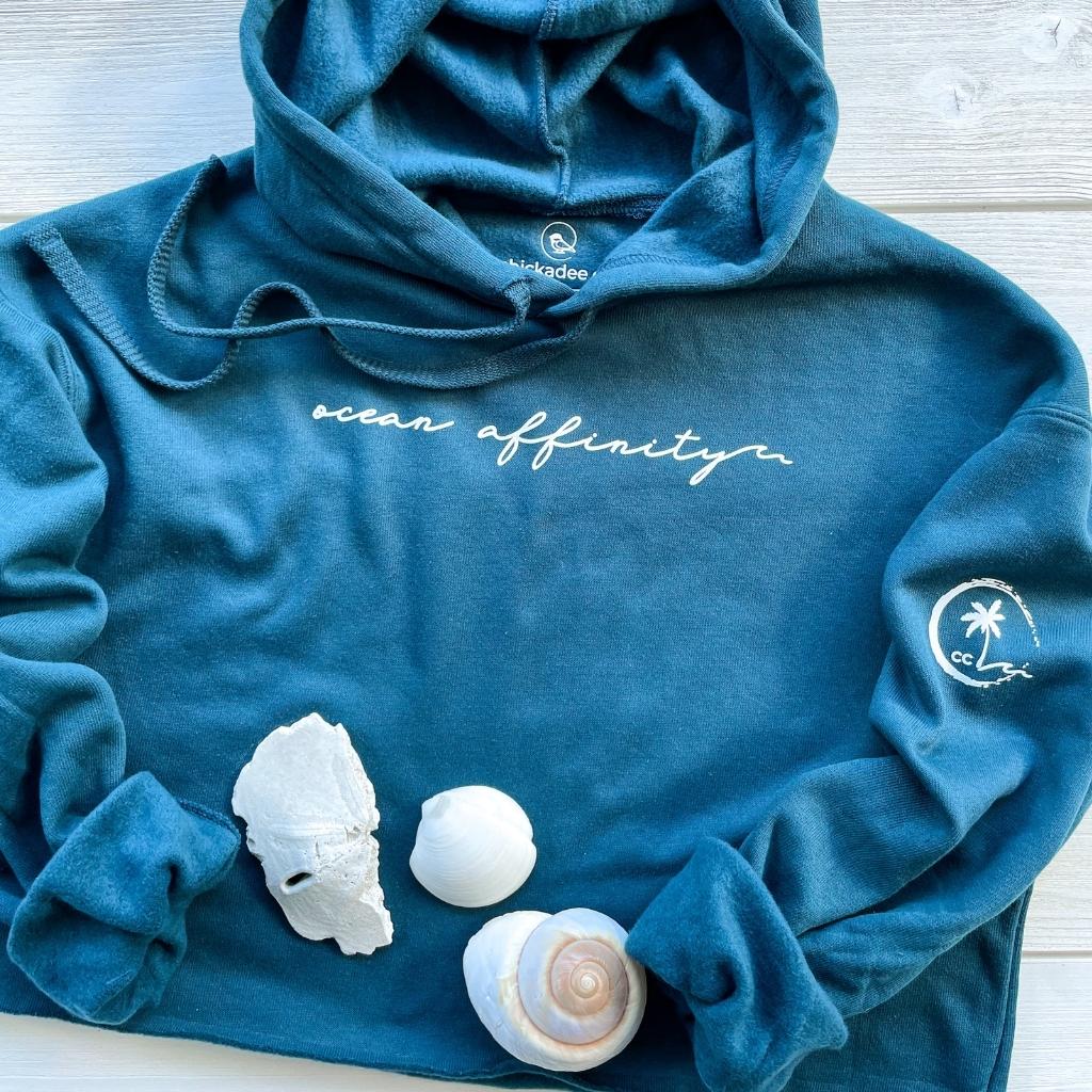 ocean affinity script white text on deep turquoise blue crop hoodie for women.  palm tree icon on sleeve.  beach apparel.  ocean lover sweatshirt.