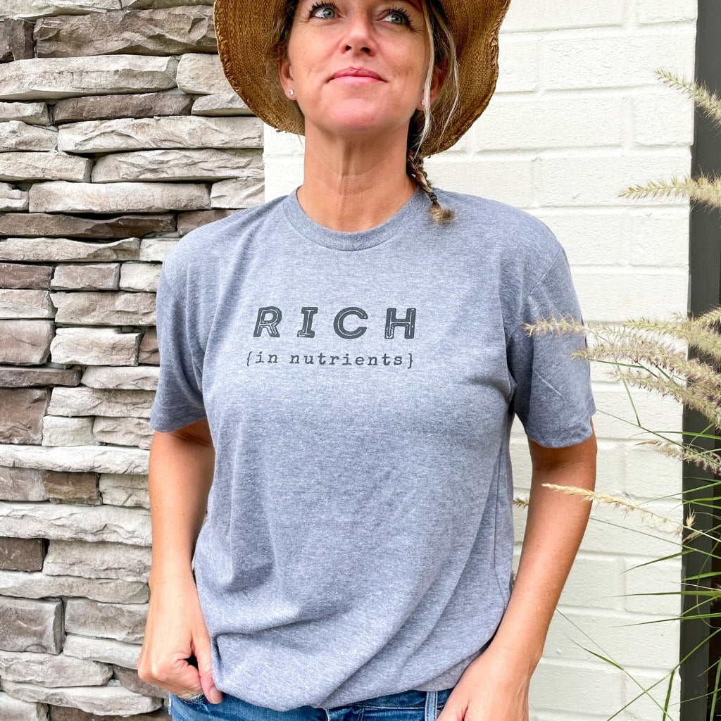 rich in nutrients black text print on vintage gray unisex tshirt.  woman wearing intentional wellness apparel.  rich in nutrients saying with black ink carrot image on sleeve.  wellness tshirt.