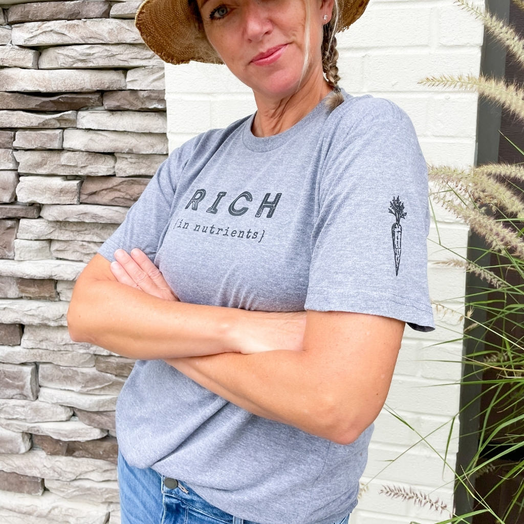 rich in nutrients black text print on vintage gray unisex tshirt.  woman wearing intentional wellness apparel.  rich in nutrients saying with black ink carrot image on sleeve.  wellness tshirt.