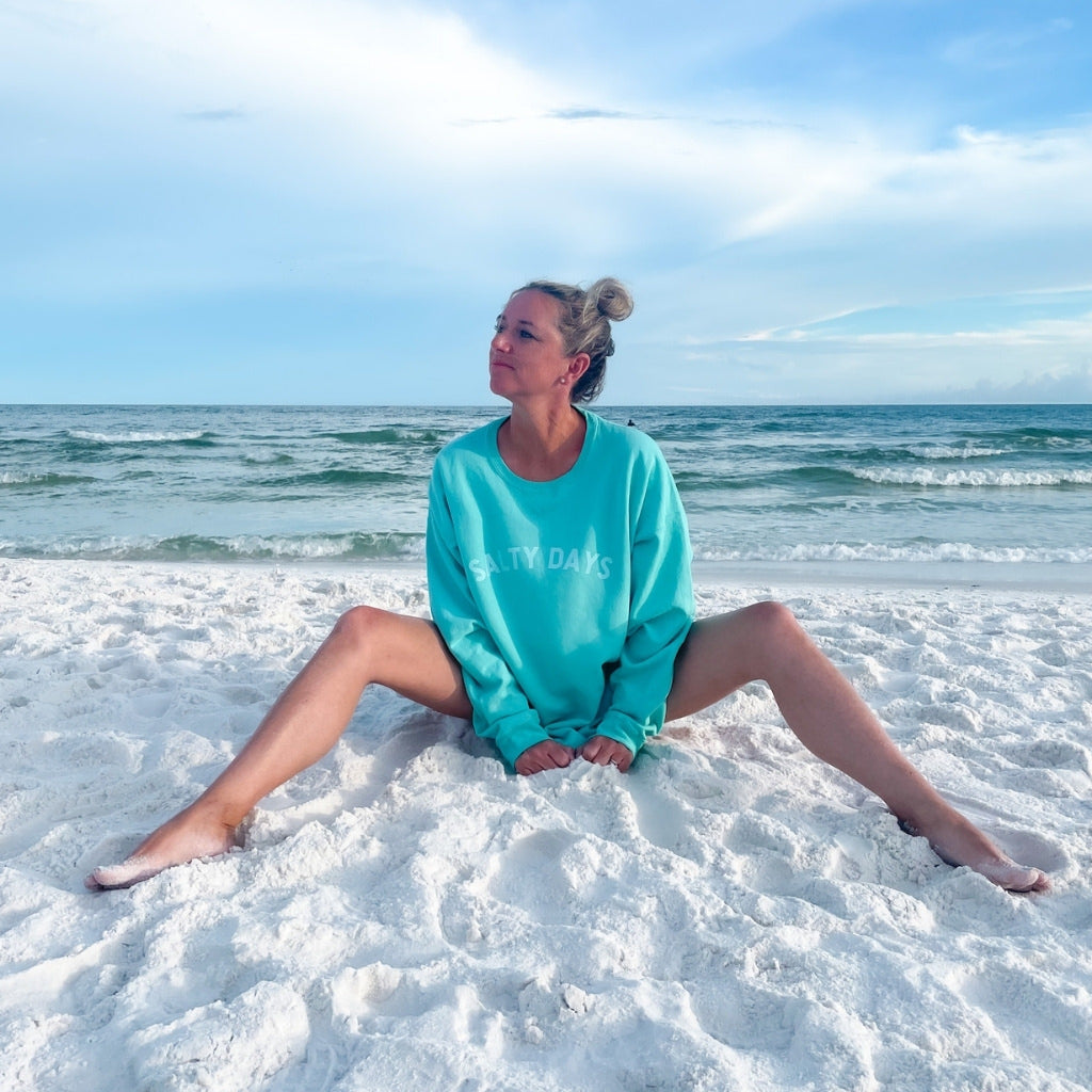 salty days simple text on aqua blue oversized cotton sweatshirt.  beach sweatshirt with woman sitting on the beach near the ocean.  beach trip clothing.