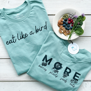 100% organic cotton tshirt in a light aqua color.  chickadee co. wellness tshirts.  womens tshirt laying flat on white boards showing the front of the tshirt printed with 'eat like a bird' and 'MORE berries, plants, seeds, nuts' on the back of the tshirt.  a small plate of healthy blueberries, cranberries, plants, nuts & seeds is shown. 