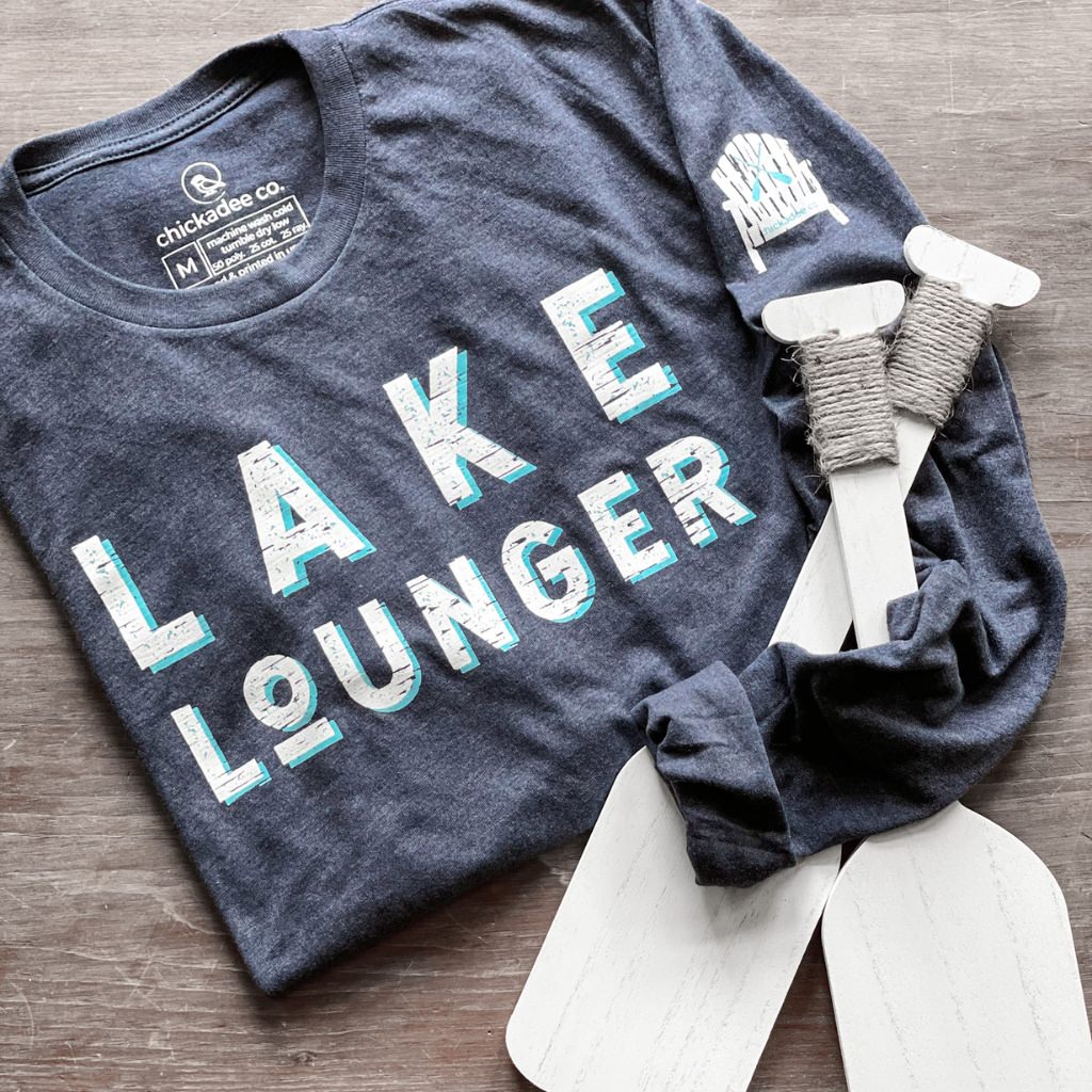 super soft womens long sleeve lake tshirt that says LAKE LOUNGER on the front in screen printed white & blue inks on a navy shirt.  cottage adirondak chair is printed on the left sleeve in white with blue oars and the chickadee co. brand name.