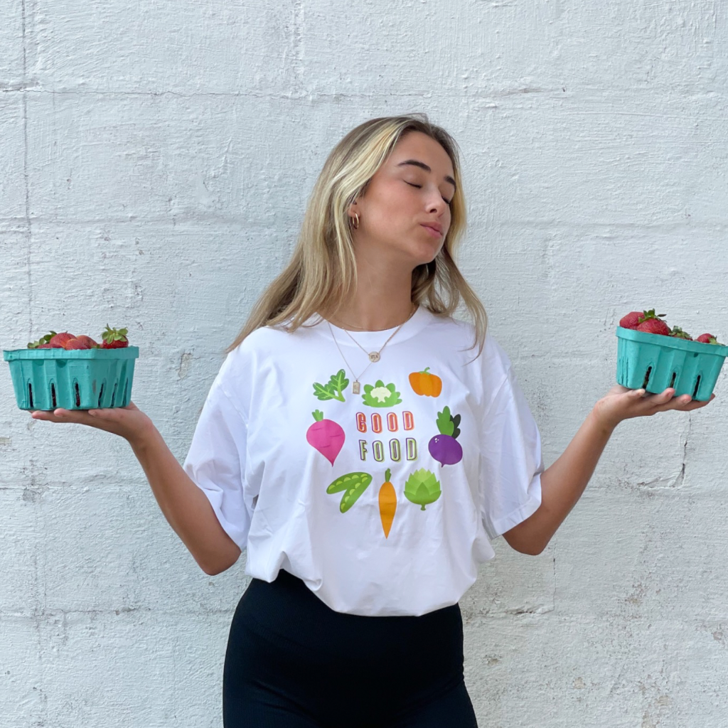 woman wearing a 100% organic cotton white tshirt.  GOOD FOOD is printed on the front in a bright colored ink shaped like a heart graphic made of vegetables.  she's holding fresh strawberries from the farmers market. 