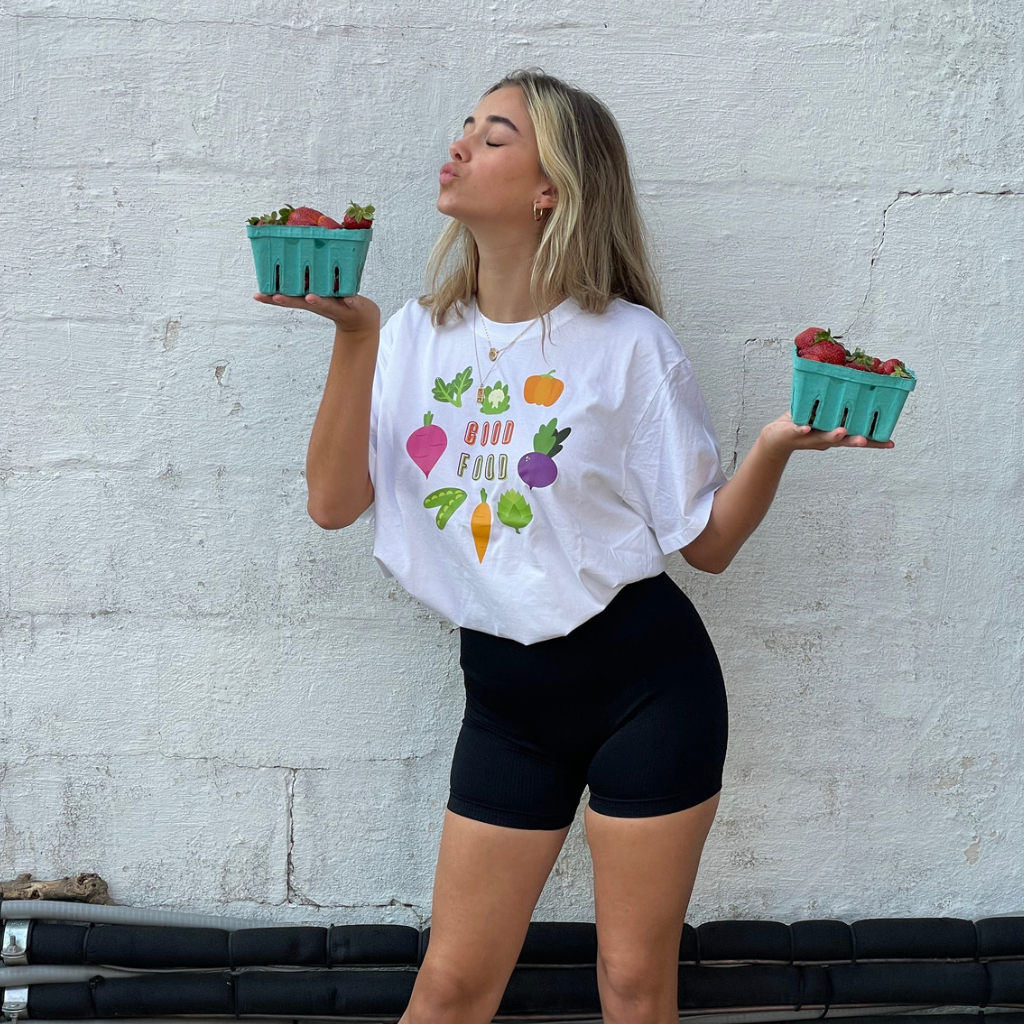 woman wearing a 100% organic cotton white tshirt.  GOOD FOOD is printed on the front in a bright colored ink shaped like a heart graphic made of vegetables.  she's holding fresh strawberries from the farmers market. 
