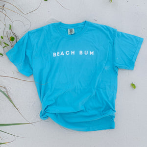 comfort colors tshirt in lagoon color laying on the sand with the word BEACH BUM printed on the chest.