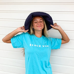 woman wearing a lagoon comfort colors BEACH BUM tshirt.  happy girl wearing a sunhat & closing her eyes with glee.