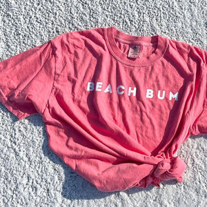 comfort colors tshirt in watermelon color laying on the sand with the word BEACH BUM printed on the chest.