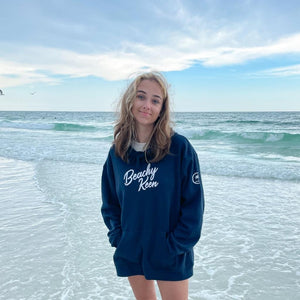 woman wearing chickadee co. Beachy Keen hoodie with shorts, standing in the turquoise blue ocean along the shore in florida gulf coast.  beach hoodie in navy.