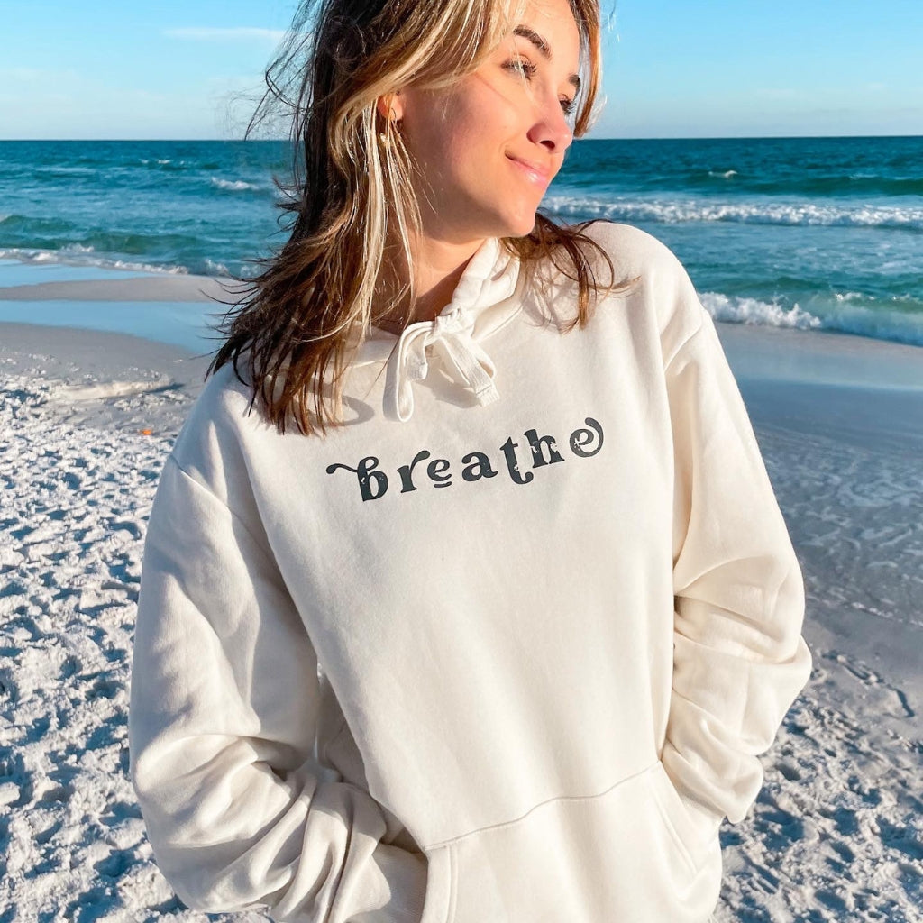 breathe.  woman wearing a cozy creamy white colored hoodie at the beach.  text simply says 'breathe' in a dark gray ink. 