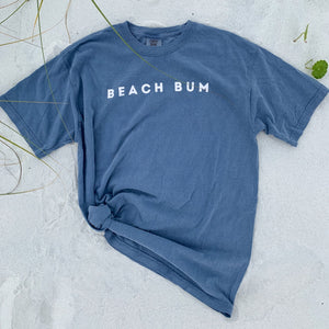 comfort colors tshirt in denim color laying on the sand with the word BEACH BUM printed on the chest.