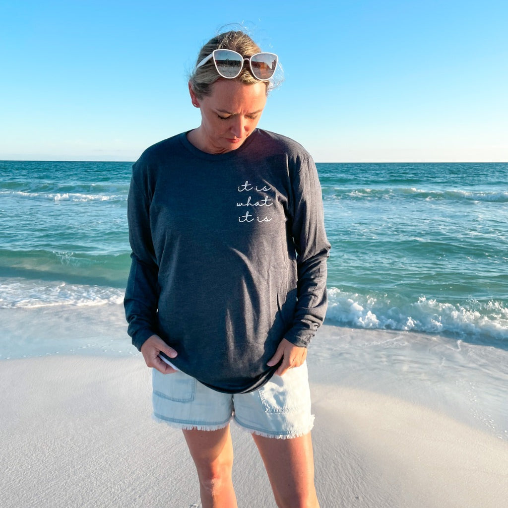 woman on the beach by the ocean wearing a navy long sleeve tshirt that says 'it is what it is' in script white text on the left chest.  inspirational clothing for women.