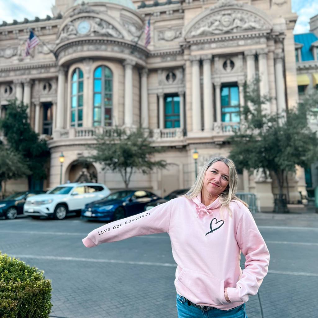 woman wearing a light pink super soft hoodie with the words 'love one another' on the right sleeve and a hand drawn heart on the left chest in black ink.  wellness hoodie for mental health and sharing kindness.  intentional hoodies for women.