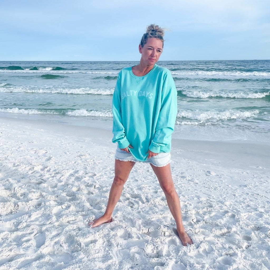 salty days simple text on aqua blue oversized cotton sweatshirt.  beach sweatshirt with woman sitting on the beach near the ocean.  beach trip clothing.