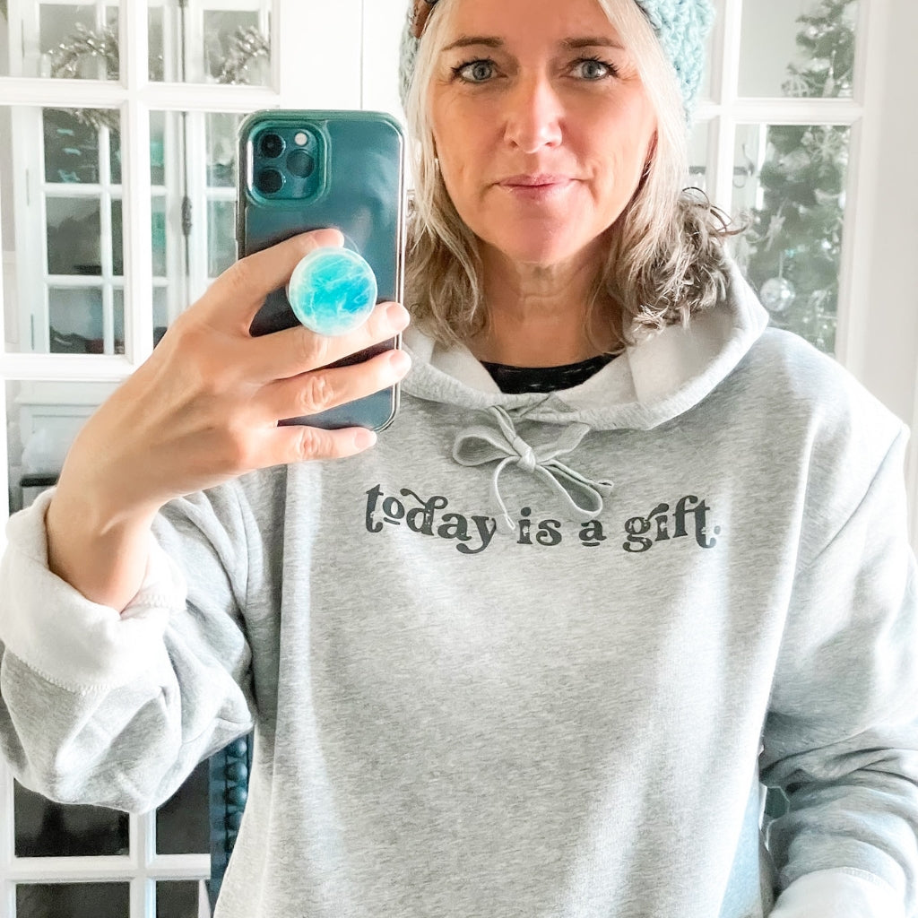 today is a gift, printed in charcoal gray ink on a heathered light gray hoodie.  unisex hoodie with screen printed image text saying today is a gift.  woman standing in front of the mirror taking a photo of herself wearing the today is a gift hoodie.  uplifting hoodie.  