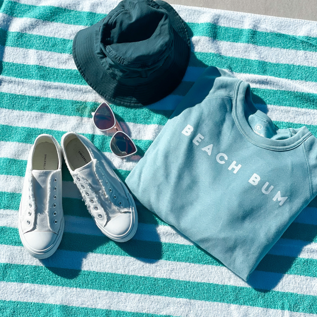 BEACH BUM sweatshirt in aqua blue laying on a beach towel with a sunhat, sunglasses & sneakers.