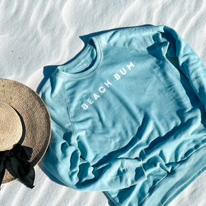 womens beach sweatshirt in aqua blue.  BEACH BUM sweatshirt laying on the white sand with a straw hat.